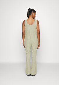 Macacão sem mangas, verde claro, com decote arredondado nas costas e pernas largas. Tecido texturizado com um padrão subtil. Calçado preto visível.