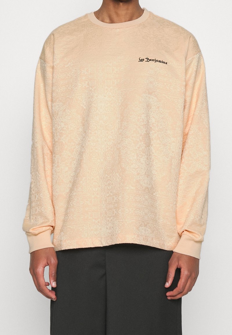 Personne portant un sweatshirt ample à motifs pêche clair avec le texte "Les Benjamins" sur la poitrine, associé à un pantalon sombre, debout devant un fond uni.