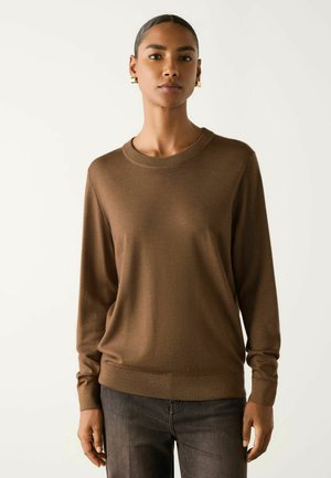 Kvinde med håret sat tilbage iført guldringeøreringe, en brun langærmet trøje og mørkegrå jeans, stående mod en ensartet lys baggrund.