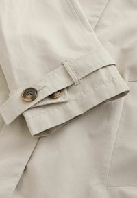 Beige trenchcoatmouw met twee bruine knopen, een gestikte lus en een gladde textuur, met een gestructureerd ontwerp en strakke lijnen.