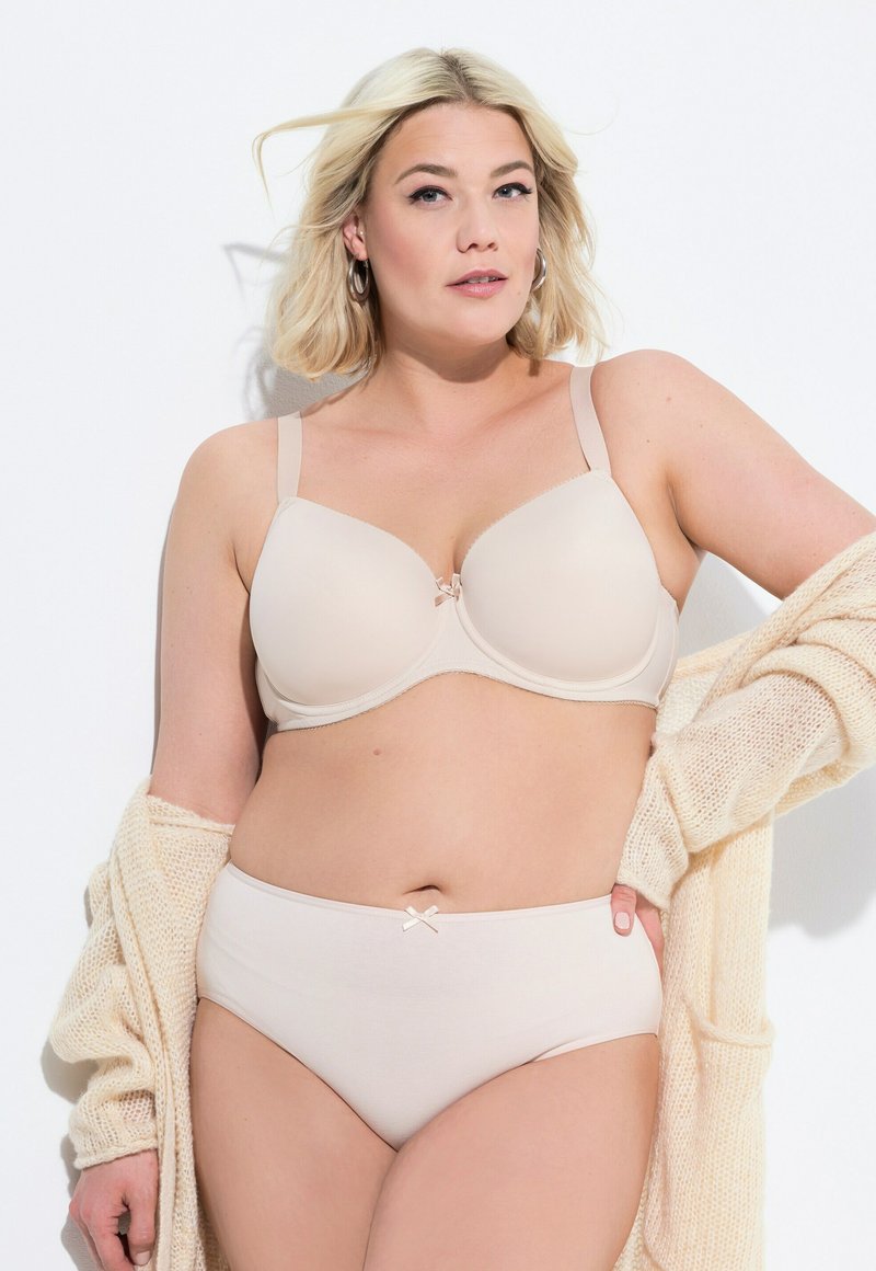 Reggiseno beige sagomato con spalline regolabili, un fiocco decorativo al centro; mutandine alte coordinate con un dettaglio di fiocco simile, cardigan testurizzato.