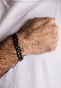Juodas grandinėlės apyrankė, pagaminta iš glotnaus, matinio medžiagos, su tarpusavyje susietais gūžiais, dėvima ant riešo priešais baltą audinį.