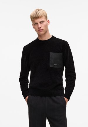 Maglione nero con colletto rotondo, polsini e orlo a costina, dotato di una tasca in tessuto nero sul lato sinistro. Texture morbida.