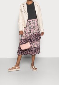Mujer vistiendo una chaqueta crema, una blusa negra, una falda midi floral rosa y azul marino, sandalias de cuerda crema, sosteniendo un pequeño bolso rosa.