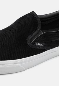 Melnas korduroja slip-on kedas ar baltu gumijas zoli, noapaļotu purngalu un elastīgām sānu akcentiem. Iekļauta Vans zīmola birka.