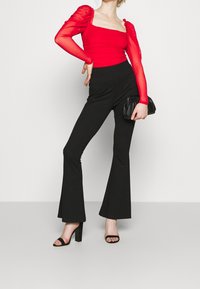 Roter taillierter Body mit transparenten langen Ärmeln, kombiniert mit schwarzen weit geschnittenen Hosen und schwarzen High-Heels. Accessoire: schwarze Clutch.