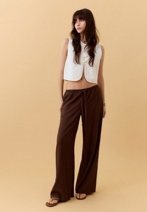 Mujer con el cabello largo y oscuro, que lleva un chaleco blanco sin mangas y corto, pantalones sueltos marrones y sandalias, de pie frente a un fondo beige.