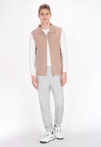Gilet in pile beige con colletto alto, indossato sopra una camicia bianca a maniche lunghe, abbinato a pantaloni della tuta grigi e scarpe da ginnastica bianche.