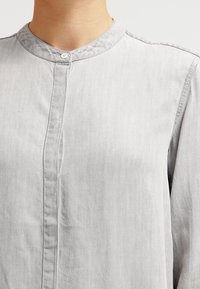 Camicia button-up grigia chiara con colletto alla mandarino, caratterizzata da una texture liscia e un design semplice della patta frontale.