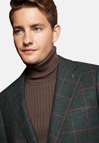 Męska wełniana marynarka w ciemnozielonym kolorze z brązowym wzorem w kratę, zestawiona z brązowym golfem o prążkowanej teksturze. Posiada przypinkę na klapie oraz detale kieszeni.