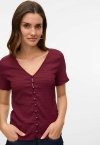 Blusa de botón marrón con cuello en V, mangas cortas y tejido texturizado. Seis botones decorativos en el frente, diseño de corte casual.