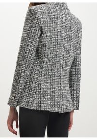 Femme portant un blazer en tweed à carreaux noir et blanc avec des boutons argentés aux poignets, associé à un pantalon noir, vue de dos.