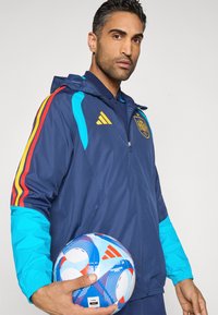 Modrý vetrovka Adidas s tyrkysovými a červenými akcentmi, so zapínaním na zips po celej dĺžke, kapucňou a emblémom. Drží modrú a červenú futbalovú loptu s vzorom.