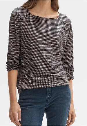 Femme portant un chemisier à manches longues, sombre à motifs, avec les manches retroussées et un jean bleu foncé, debout devant un fond uni.