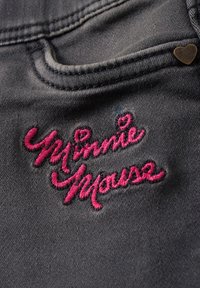 Tissu en denim gris foncé avec une poche avant, décoré d’un texte rose brodé « Minnie Mouse » et d’un petit rivet métallique en forme de cœur.