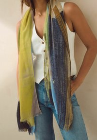 Femme portant un haut blanc sans manches, un jean bleu et une longue écharpe multicolore avec des motifs jaunes, bleu marine et beiges.