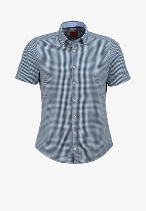 Homme portant une veste en cuir marron, une chemise à carreaux bleue, un pantalon gris clair et des chaussures en cuir marron, debout avec une main dans la poche.