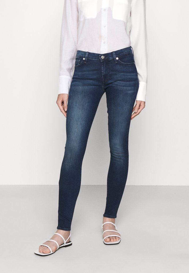 Jeans skinny azul oscuro con una textura de denim suave, diseño de cinco bolsillos y una cinturilla de altura media. Combinados con una camisa de botones clara.
