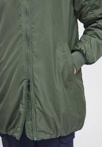 Giacca a vento verde oliva con chiusura a zip, polsini elasticizzati e tasche laterali. Il tessuto ha una consistenza liscia e dettagli di design minimi.