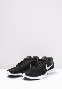 Pár černobílých běžeckých bot Nike s bílým logem ve tvaru háčku, černými tkaničkami a bílou podrážkou na bílém povrchu.