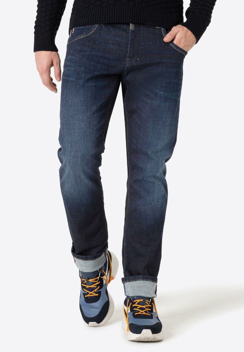 Donkerblauwe denim jeans met een getailleerde pasvorm en lichte vervaging, gecombineerd met multicolour sneakers met oranje en grijze accenten.