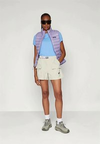 Naine mustade prillidega, heledast lillast pufferovest, sinisest T-särgist, beežidest cargo-lühikestest pükstest, valgetest sokkidest ja hallidest matkasaapadest, seisab käsi taskus.