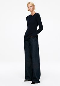 Marineblaue gerippte Langarmbluse mit taillierter Silhouette, kombiniert mit weit geschnittenen, dunklen Jeans. Schwarze spitz zulaufende Schuhe runden das Outfit ab.