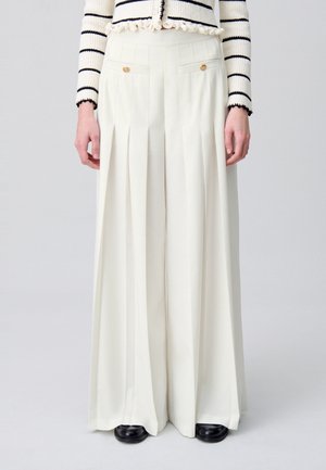 Witte broek met wijde pijpen, voorplooien en gouden knoopaccenten, gedragen met een zwart-witte gestreepte trui en zwarte schoenen.