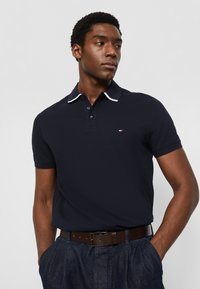 Polo blu navy con texture a maglia, bordo colletto bianco a contrasto e patta con tre bottoni; indossato con cintura marrone e pantaloni di denim.
