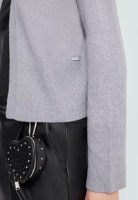 Jasnoszary dziergany kardigan z żebrowanym dołem i mankietami, zestawiony z czarnymi skórzanymi spodniami oraz małą czarną torebką w kształcie serca z ćwiekami.