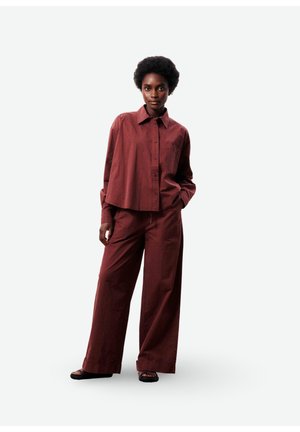 Femme aux cheveux courts et bouclés portant une chemise rouge foncé ample à boutons et un pantalon à jambes larges, debout avec une main dans la poche.