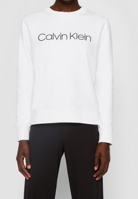 Vit sweatshirt med rund hals, ribbade muddar och fåll, med svart "Calvin Klein"-logotyp på framsidan. Stylad med svarta byxor.