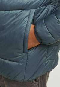 Puffervest i mørk teal med polstret tekstur, sidelomme og elastiske manchetter for varme og komfort.