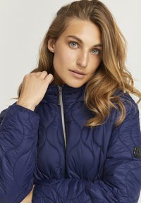 Giacca trapuntata blu navy con colletto alto e zip frontale. Presenta una texture liscia e un patch rotondo con logo sulla manica sinistra.