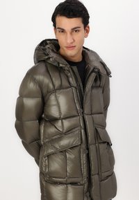 Veste rembourrée vert olive avec capuche, dotée d'une fermeture éclair à l'avant, de boutons-pression et de multiples poches ; tissu brillant et texturé.