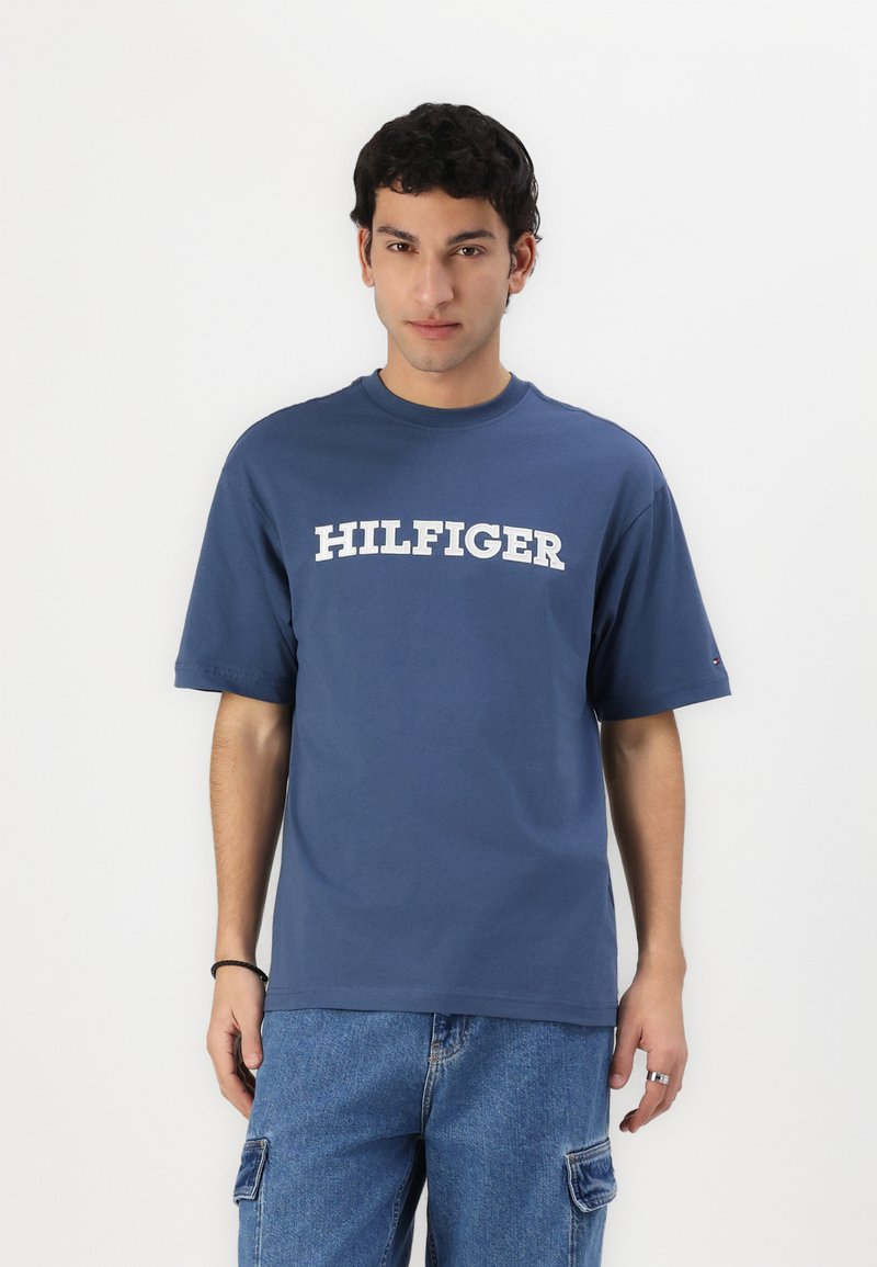 Musta puuvillapaita, jossa on lyhyet hihat ja pyöreä kaula-aukkko, jossa on suuri valkoinen "HILFIGER" -teksti rinnassa.