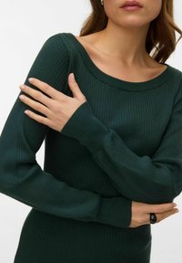 Tmavě zelený žebrovaný pletený svetřík se širokým výstřihem a dlouhými rukávy, s přiléhavým střihem a texturovanou látkou. Na ruce černý prsten.