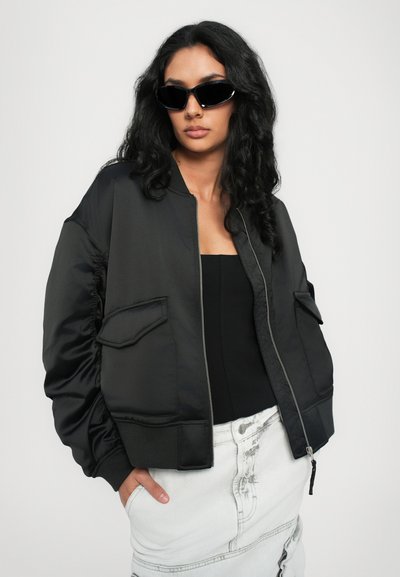 Veste bomber noire en tissu satiné, avec des poignets côtelés et deux poches avant. Portée par-dessus un top noir et une jupe en denim blanche. Lunettes de soleil noires.