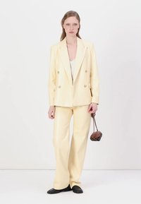 Blazer doppiopetto di colore crema con pantaloni a gamba larga realizzati in materiale lucido, abbinati a una borsa scura e scarpe nere.