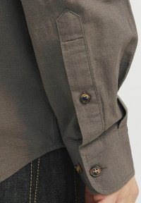 Puño de camisa de lino marrón con dos botones marrones, que presenta un detalle de costura triangular en la manga. Tejido oscuro debajo con costura amarilla.