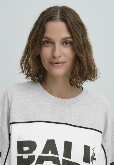 Šedá a biela polovičná košeľa s potlačou "BALI" vo výrazných čiernych písmenách na hrudi. Má okrúhly výstrih a kontrastné lemovanie.