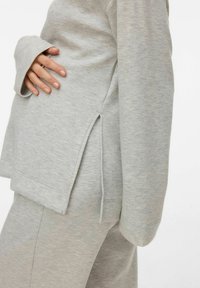 Svijetlosiva majica s dugim rukavima, bočnim prorezima na rubu i mekanim teksturiranim tkanjem, u kombinaciji s odgovarajućim sivim hlačama.