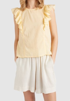 Gelbes Kurzarm-Top mit Rüschendetails entlang der Schultern, kombiniert mit hellbeigen, locker sitzenden Shorts. Weicher, strukturierter Stoff.