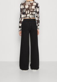 Svarta vida jeans i mjuk bomullstextur, kombinerat med en genomskinlig långärmad topp med ett abstrakt vitt mönster och volangkanter.