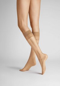 Beige knielange Strümpfe mit glatter Textur und transparenter Optik, ausgestattet mit elastischen Bändern am oberen Ende, die ein dezentes Logo tragen.