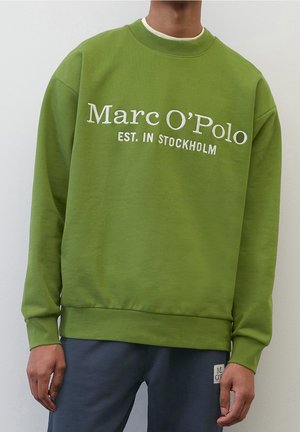 Osoba ubrana w zieloną bluzę z napisem "Marc O’Polo EST. IN STOCKHOLM" oraz ciemne spodnie na jednolitym białym tle.