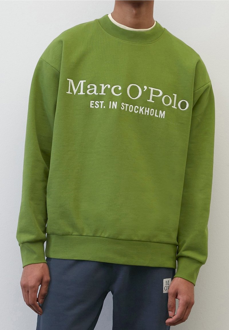 Personne portant un sweat-shirt vert avec le texte "Marc O’Polo EST. À STOCKHOLM" et un pantalon foncé sur un fond blanc uni.