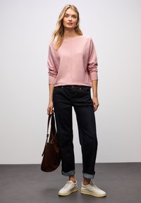 Helles rosa Strickpullover mit rundem Ausschnitt, kombiniert mit dunkelblauen Jeans mit umgeschlagenem Saum und cremefarbenen Sneakers. Eine braune Schultertasche ist ebenfalls enthalten.