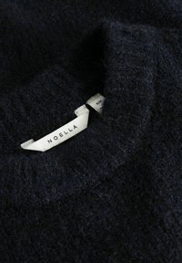 Pull duveteux bleu marine foncé avec col côtelé montrant une étiquette blanche avec le nom de la marque "NOELLA".