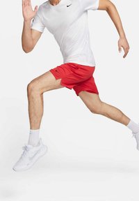 Corridore a metà corsa che indossa una maglietta bianca Nike, pantaloni corti rossi Nike, calzini bianchi e scarpe da ginnastica bianche Nike su uno sfondo bianco semplice.